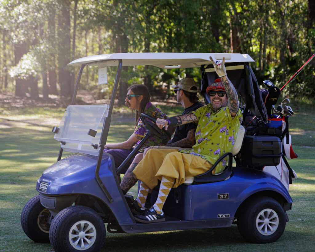 Photo of three golf players from Golf Wars 2024 riding in a golf cart with golf clubs strapped to the rear of the cart. The driver is flashing a rocking horns hand signal to the camera while smiling.