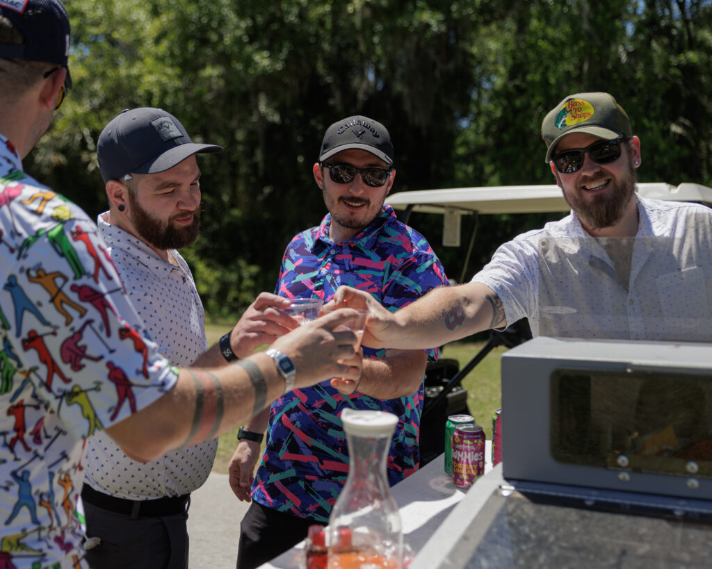 Photo of 4 golf players clinking drinking glasses full of booze they'd just bought from the drink cart at Golf Wars 2024. All players are wearing baseball hats and sunglasses.