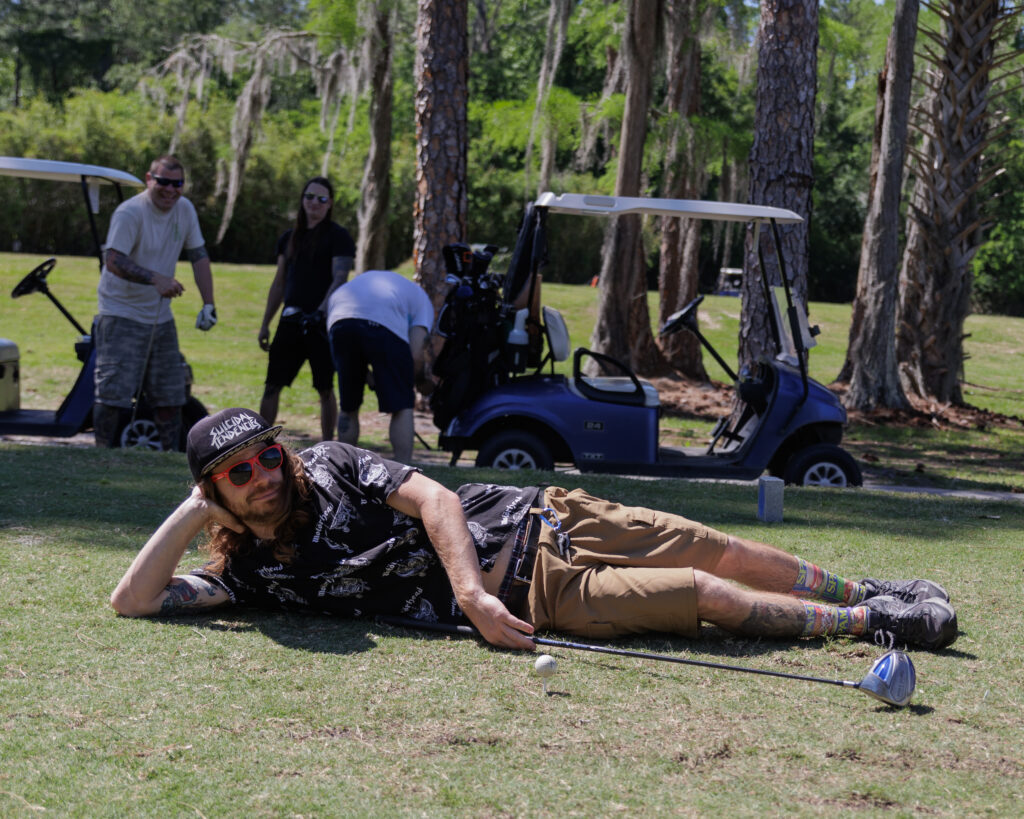 Photo of a golf player from Golf Wars 2024 laying on the driving range posing for the camera. The other three members of his team can be seen in the background of the photo organizing their golf clubs on the back of the golf carts.