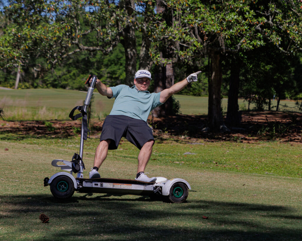 Photo of a golfer from Golf Wars 2024 riding on a motorized scooter. The player is leaning back and pointing towards the sky in a victory pose. The scooter is riding on only 3 of the 4 wheels.